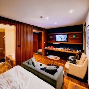 Sala de estar de hotel con sofás grises, televisión de pantalla plana y mobiliario en madera clara