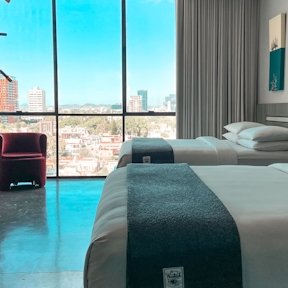 Dormitorio de hotel con cama doble, manta decorativa azul y gran ventana con vista urbana